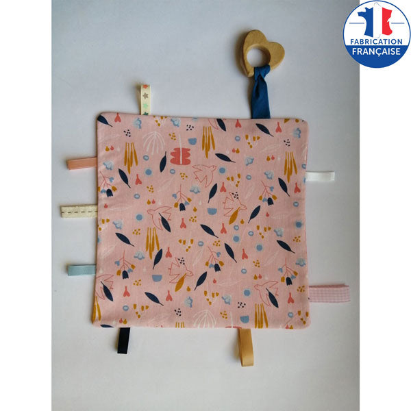 Un côté en tissu 100 % coton imprimé, l’autre en tissu minky coloré. Tout autour, des rubans aux textures, tailles et couleurs différentes. Un anneau en bois non traité que bébé pourra mordiller et attraper.